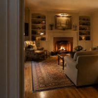 A white paddle dimmer on a dark wall beside a doorway, with a warmly lit living room beyond it: a lit fireplace, table lamps and a picture light over a painting.