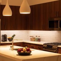 A kitchen at night with warm under-cabinet lighting along the counters and two pendant lights over the counter run, in a room of dark walnut cabinets and a white quartz island.