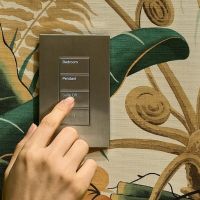 A hand pressing a bronze wall keypad with engraved buttons labeled Bedroom and Pendant.