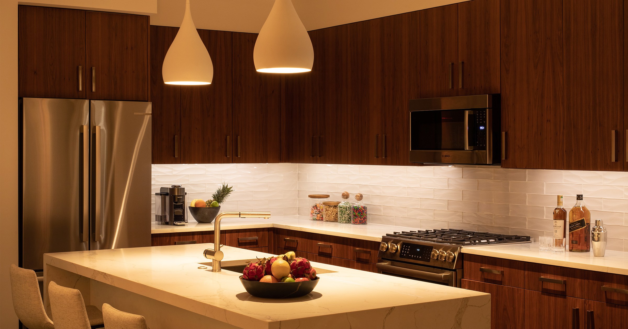 A kitchen at night with warm under-cabinet lighting along the counters and two pendant lights over the counter run, in a room of dark walnut cabinets and a white quartz island.