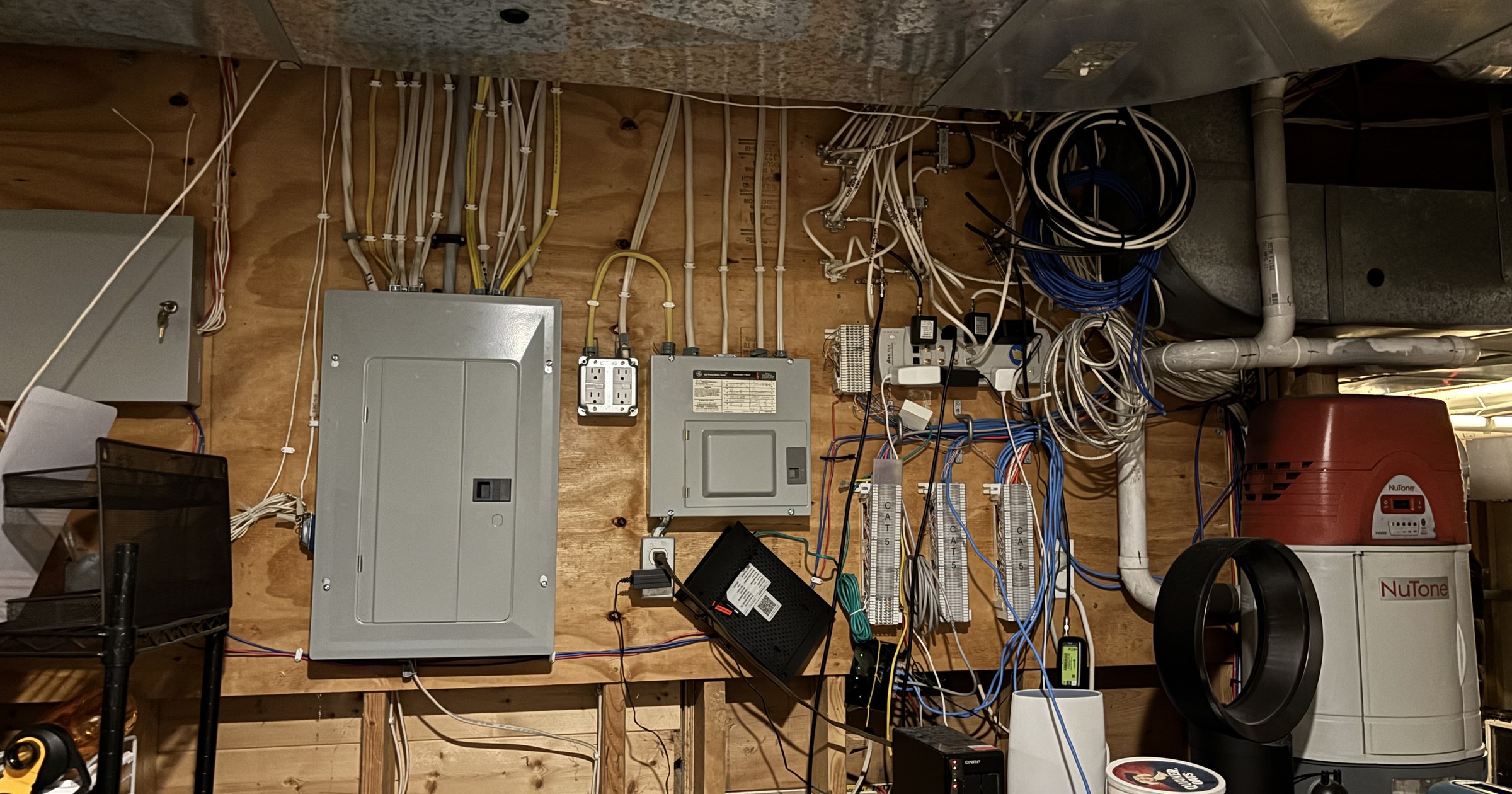 A utility room wall with an electrical panel, cable modem, patch panels, tangled network cabling, and equipment added over several years by different owners.