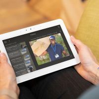 A woman holds a tablet in her hands as she looks at a security camera feed of a package delivery.