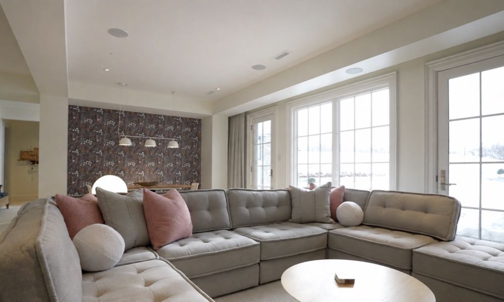 <span class="uk-h4">A living room with a sectional sofa and three flush in-ceiling speakers, with snow visible through the windows.</span>