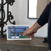 Person tapping a touchscreen control panel with Control4 smart home interface.