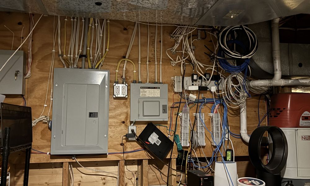 <span class="uk-h4">A utility room wall with an electrical panel, cable modem, patch panels, tangled network cabling, and equipment added over several years by different owners.</span>