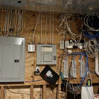 A utility room wall with an electrical panel, cable modem, patch panels, tangled network cabling, and equipment added over several years by different owners.